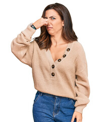Young brunette woman wearing casual winter sweater smelling something stinky and disgusting, intolerable smell, holding breath with fingers on nose. bad smell - obrazy, fototapety, plakaty