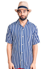 Young handsome man with beard wearing summer hat and striped shirt smiling looking to the side and staring away thinking.
