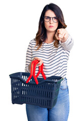 Beautiful young brunette woman holding supermarket shopping basket pointing with finger to the camera and to you, confident gesture looking serious