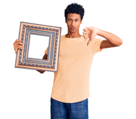 Young african american man holding empty frame with angry face, negative sign showing dislike with thumbs down, rejection concept