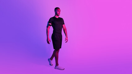 Fototapeta na wymiar Sporty African Man With Phone Armband Walking On Purple Background