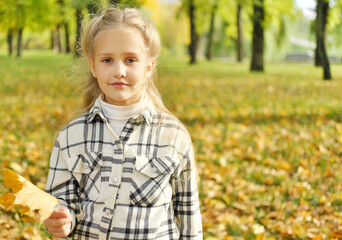 A little preschool blonde girl holds an autumn leaf in her hands, smiles and looks at the camera