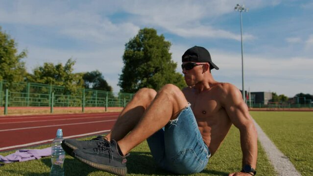 Bodybuilder with naked torso has intense training in outdoor sports field and does abs crunch over a bottle