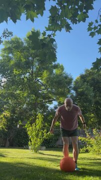 senior, athletic man is exercising in a backyard with a heavy slam ball, green summer scenery