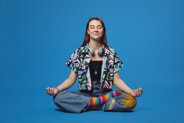 Calm young girl with headphones wearing stylish colorful clothes, meditating with eyes closed while practicing mindfulness, sitting on floor in lotus pose, isolated over blue background - obrazy, fototapety, plakaty