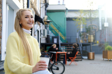 A blonde girl in a yellow sweater and jeans sits on a bench with a cup of takeaway coffee. The girl looks away and smiles