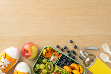 Nutritious lunchbox arrangement in school setting. Top view of plastic container with fresh produce, sandwich, assorted berries, water bottle, sneakers, wooden desk backdrop, space for text or ad