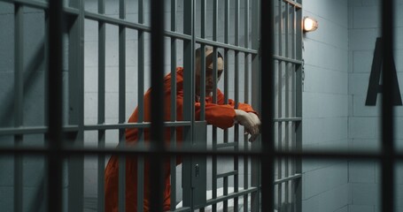 Elderly prisoner in orange uniform stretches fingers, leans on metal bars. Criminal serves...