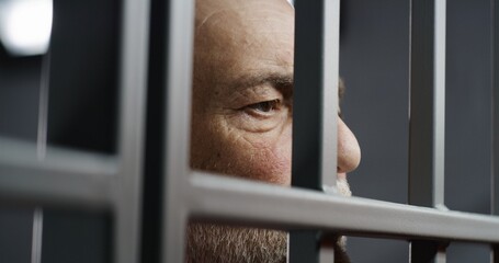 Face close up of depressed elderly prisoner standing behind metal bars in prison cell. Criminal or murderer serves imprisonment term for crime. Inmate in jail or detention center. Justice system.