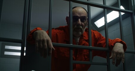 Elderly prisoner in orange uniform holds hands on metal bars in jail. Guilty criminal serves...