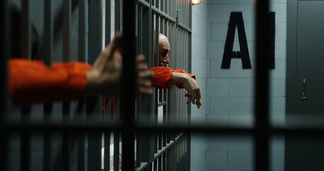 Prisoners in orange uniforms lean on metal bars in prison cells. Guilty criminals serve...