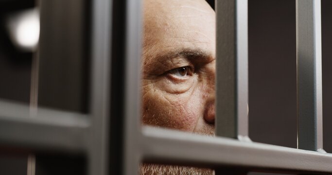 Face close up of depressed elderly prisoner standing behind metal bars in prison cell. Criminal or murderer serves imprisonment term for crime. Inmate in jail or detention center. Justice system.