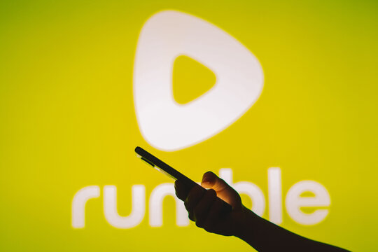 July 12, 2023, Brazil. In this photo illustration the Rumble logo seen in the background of a silhouette of a woman holding a mobile phone.