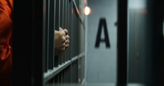 Close up of prisoner in orange uniform holding metal bars, standing in prison cell. Guilty criminal or killer serves imprisonment term for crime. Inmate in jail or detention center. Justice system.