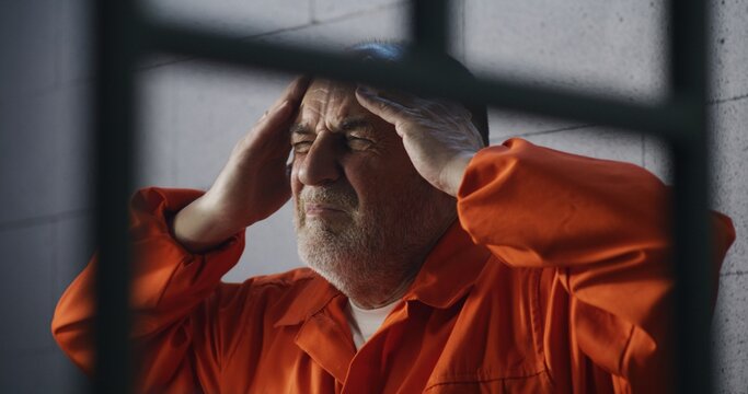 Senior criminal in orange uniform sits on prison bed and dreams about freedom. Prisoner serves imprisonment term in jail cell. Depressed inmate in detention center or correctional facility.
