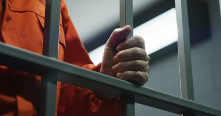 Close up of prisoner in orange uniform holding metal bars, standing in prison cell. Guilty criminal...