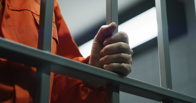 Close up of prisoner in orange uniform holding metal bars, standing in prison cell. Guilty criminal or killer serves imprisonment term for crime. Inmate in jail or detention center. Justice system.