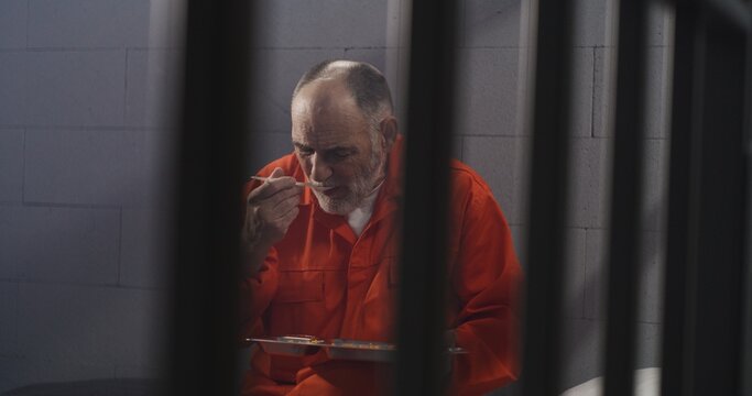 Elderly prisoner in orange uniform sits in prison cell. Prison guard gives him tray of food through metal bars. Guilty criminal serves imprisonment term for crime. Inmate in jail or detention center.