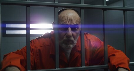 Elderly prisoner in orange uniform holds hands on metal bars, looks at camera. Criminal serves term of imprisonment in prison cell. Inmate stands behind bars in jail or detention center. Portrait.