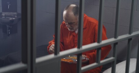 Elderly prisoner in orange uniform sits in prison cell, eats food. Criminal serves imprisonment term for crime. Conditions in jail or correctional facility. View from jail cell through metal bars.