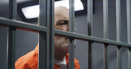 Face close up of elderly prisoner in orange uniform standing behind metal bars in prison cell....