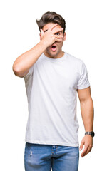 Young handsome man wearing white t-shirt over isolated background peeking in shock covering face and eyes with hand, looking through fingers with embarrassed expression.