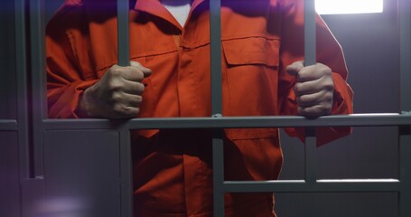 Elderly prisoner in orange uniform holds metal bars, stands in prison cell. Guilty criminal or...