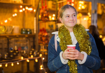 Cute brown-haired teen girl in festive mood with gold tinsel around her neck holding paper cup of...