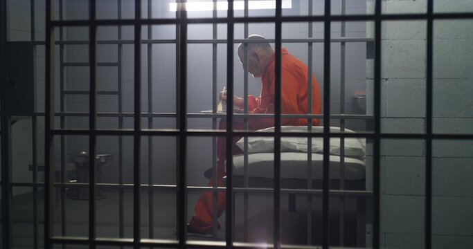 Elderly prisoner in orange uniform sits in prison cell, eats food. Criminal serves imprisonment term for crime. Conditions in jail or correctional facility. Shooting from jail cell through metal bars.