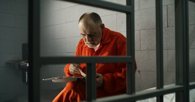 Elderly prisoner, inmate in orange uniform eats food sitting in prison cell. Criminal serves imprisonment term for crime. Jail, detention center or correctional facility. Shooting through metal bars.