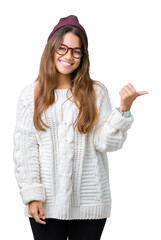 Young beautiful brunette hipster woman wearing glasses and winter hat over isolated background smiling with happy face looking and pointing to the side with thumb up.