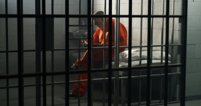 Elderly prisoner in orange uniform sits in prison cell, eats food. Criminal serves imprisonment term for crime. Conditions in jail or correctional facility. Shooting from jail cell through metal bars.