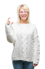 Young beautiful blonde woman wearing winter sweater and glasses over isolated background smiling and confident gesturing with hand doing size sign with fingers while looking and the camera