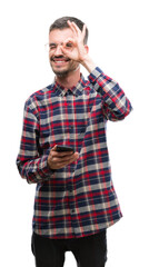 Young hipster adult man holding smartphone with happy face smiling doing ok sign with hand on eye looking through fingers