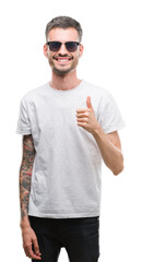 Young tattooed adult man wearing sunglasses happy with big smile doing ok sign, thumb up with fingers, excellent sign