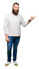 Young blond man wearing casual sweater with a big smile on face, pointing with hand and finger to the side looking at the camera.