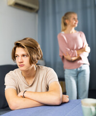 Frustrated teenage boy sitting at the table at kitchen after conflict with his mother