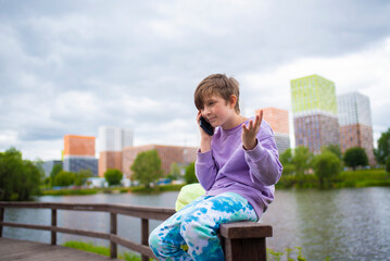 Fototapeta na wymiar emotional boy talking using a smartphone, calling his parents or friends, a schoolboy is sitting on a fence on the river embankment in the city and talking on a mobile phone.