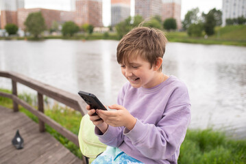 emotional child plays games on the phone, communicates with friends online, watches streams. boy sits on the embankment of the river in the city, after school.