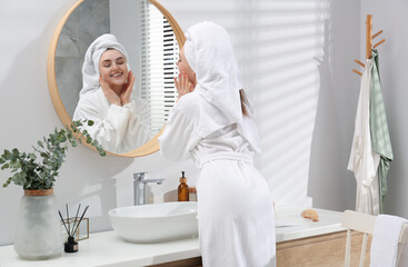 Beautiful woman in white robe applying cream near mirror in bathroom