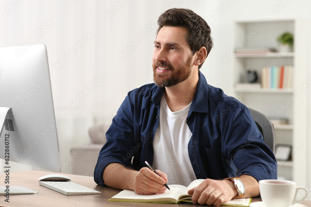 Sticker Home workplace. Happy man taking notes while working with computer at wooden desk in room