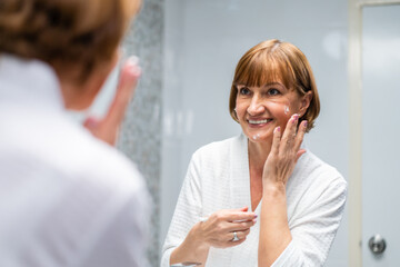 Caucasian senior woman looking at mirror then apply lotion on face. 