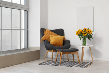 Grey armchair, footstool and vase with beautiful sunflowers near white wall