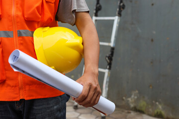 Front view of male construction worker holding blueprint and safety helmet outdoor of construction...