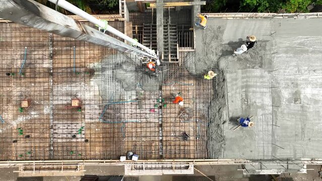 Aerial view of several workmen in hardhats pouring ready-mixed concrete after placing steel reinforcement on construction site. Foreman in blue uniform leveling floor screed