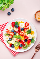 Summer fruit and berry salad with fresh strawberries, blueberries, banana, kiwi, orange and mint, pink background, top view