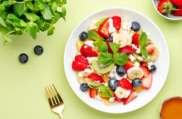 Summer fruit and berry salad with feta cheese, fresh strawberries, blueberries, banana, kiwi, orange and mint, green background, top view