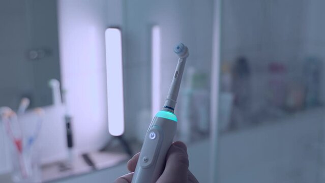 Mans hand turned on or off sonic white teethbrush with blue light against bathroom background. Dental hygiene and clean whites of the teeth. Electrical toothbrush. Power tool for cleaning mouths. 