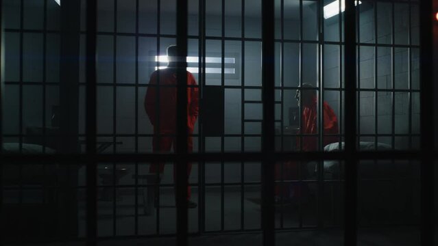 Two guilty inmates in detention center or correctional facility. Elderly criminal in orange uniform sits on jail bed and talks with cellmate. Gloomy prisoners serve imprisonment term in prison cell.