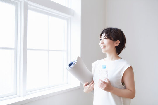 窓辺でヨガマットとペットボトルを持つ女性